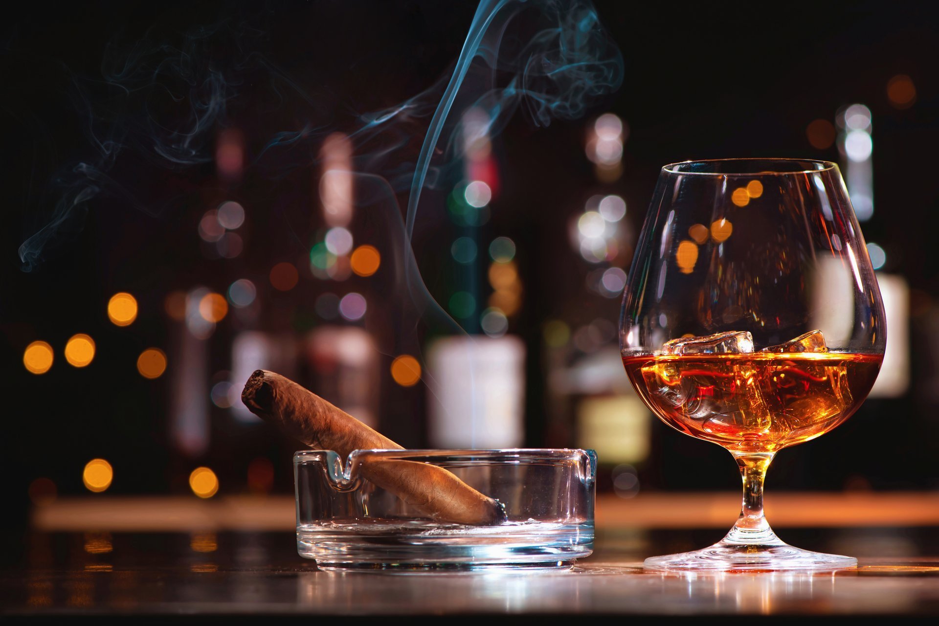 Glass of whiskey or cognac with ice cubes and smoking cigar on bar counter