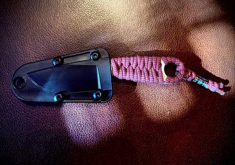 Small fixed-blade knife with black sheath and pink paracord lanyard on brown leather background