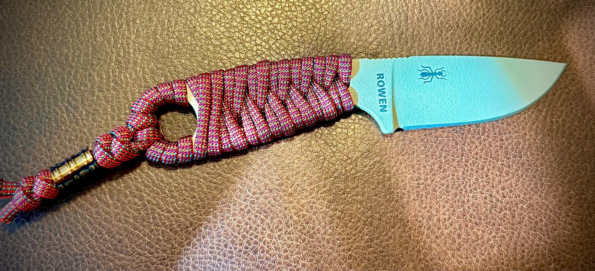 A kitchen knife with a light blue blade and a pink and black checkered fabric handle sheath on a tan surface