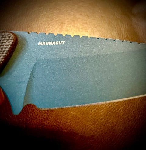 Close-up of a blue Magnacut knife blade with serrated edge resting on a brown textured surface