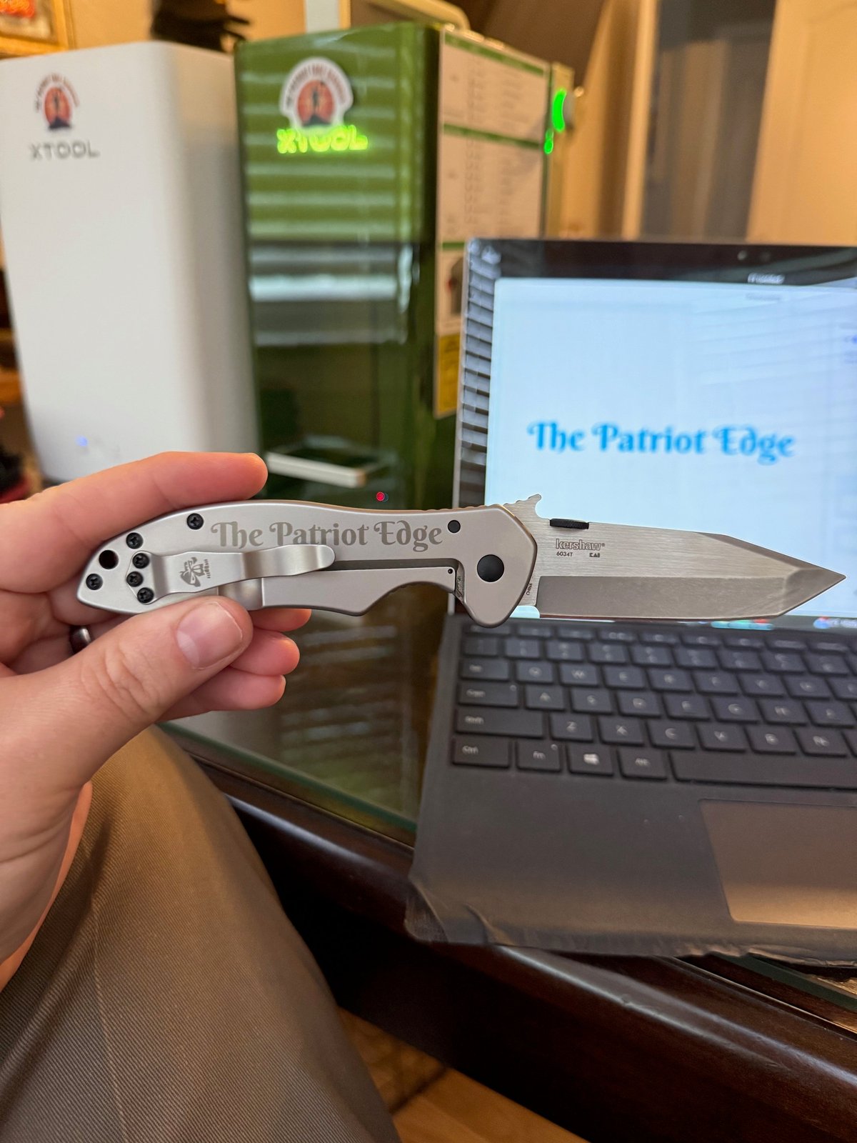 Hand holding an open pocket knife with The Patriot Edge engraved on the blade, displayed in front of a laptop showing matching text