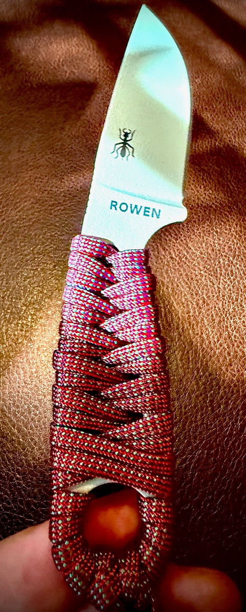 Rowen brand knife with pale blade, ant logo, and burgundy textured wrapped handle held over brown fabric