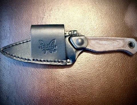 Small fixed-blade knife with tan handle and black leather sheath featuring embossed logo on tan textured background